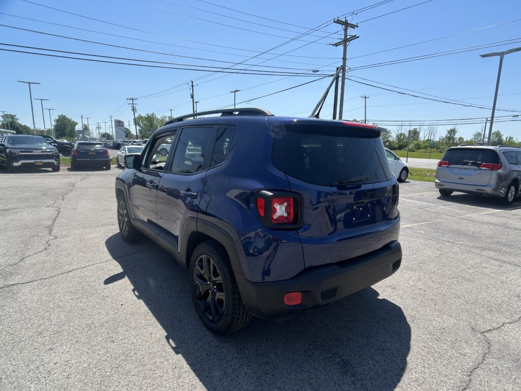 Rear three-quarter view of a blue Jeep SUV in a crowded parking lot beneath a clear blue sky, with other cars nearby and power lines overhead.