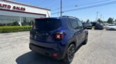 Blue Jeep SUV in a car dealership lot, rear view, with an Auto Sales building and many cars in the background on a sunny day.