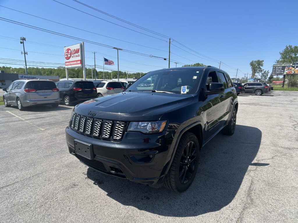 Black Jeep SUV in the foreground of a car dealership lot with other vehicles, an EZ Loan sign, and an American flag against a clear blue sky.
