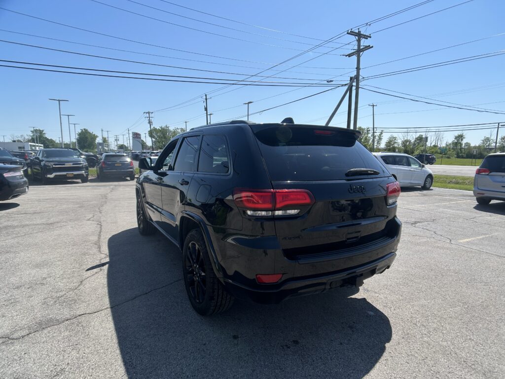 Rear three-quarter view of a black Jeep SUV parked in a busy car lot with power lines overhead and clear blue sky.