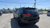 Rear three-quarter view of a black Jeep SUV parked in a busy car lot with power lines overhead and clear blue sky.