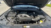 Open car hood reveals engine bay with battery, air intake, and various hoses and wires visible under bright daylight in a parking lot