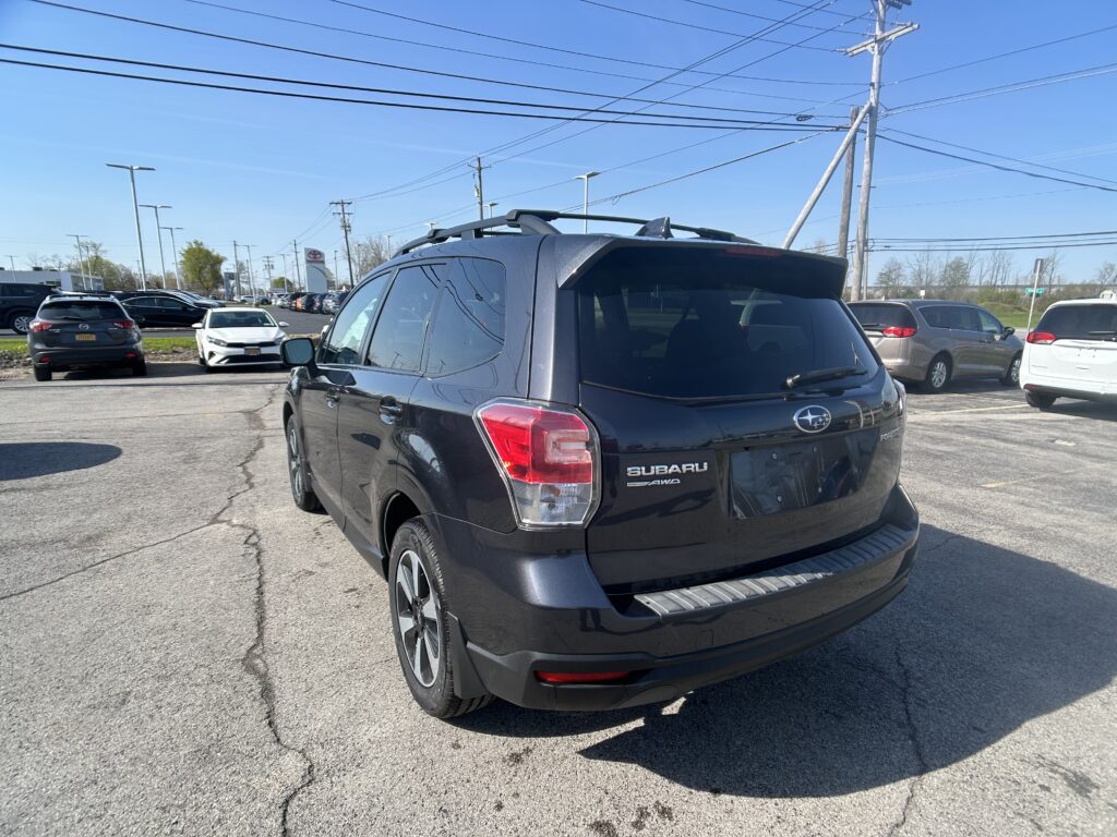 Rear view of a dark Subaru SUV with a roof rack in a busy car dealership lot, other vehicles visible in the background, power lines overhead.