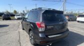 Rear view of a dark Subaru SUV with a roof rack in a busy car dealership lot, other vehicles visible in the background, power lines overhead.