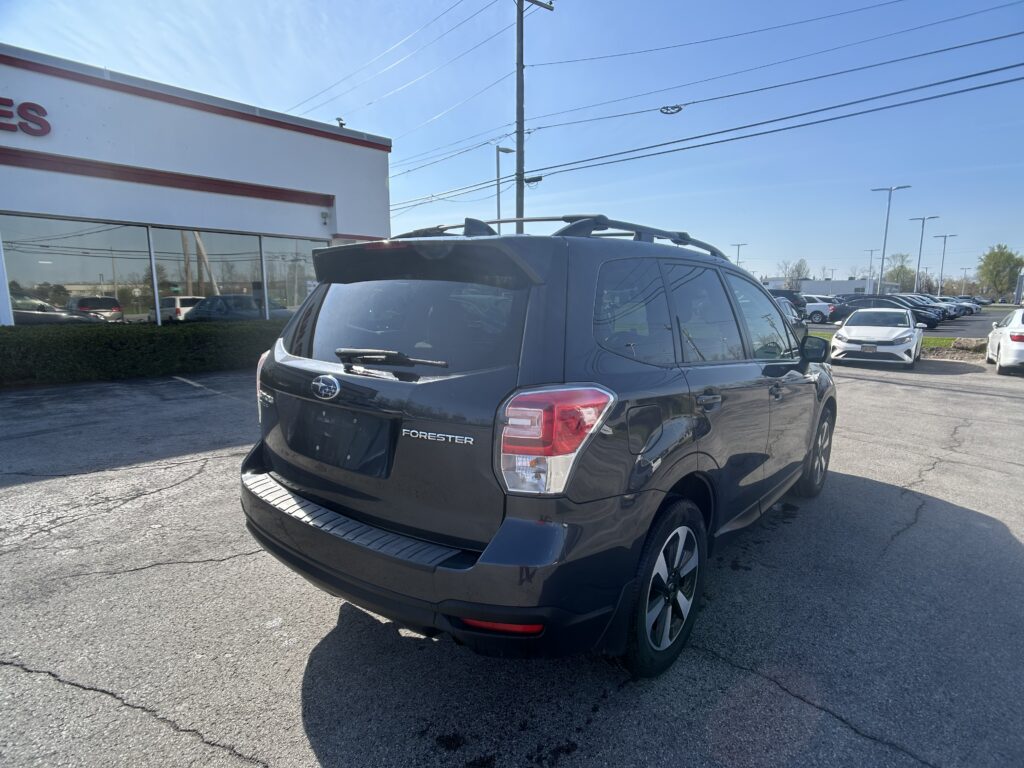 Rear three-quarter view of a black Subaru Forester SUV with roof rails, parked in a dealership lot near a white building with red trim.
