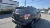 Rear three-quarter view of a black Subaru Forester SUV with roof rails, parked in a dealership lot near a white building with red trim.