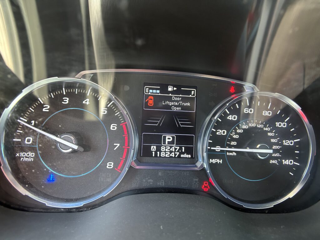 Dashboard instrument cluster with a tachometer on the left, speedometer on the right, and a center display showing Door/Liftgate Open and gear indicator (P).