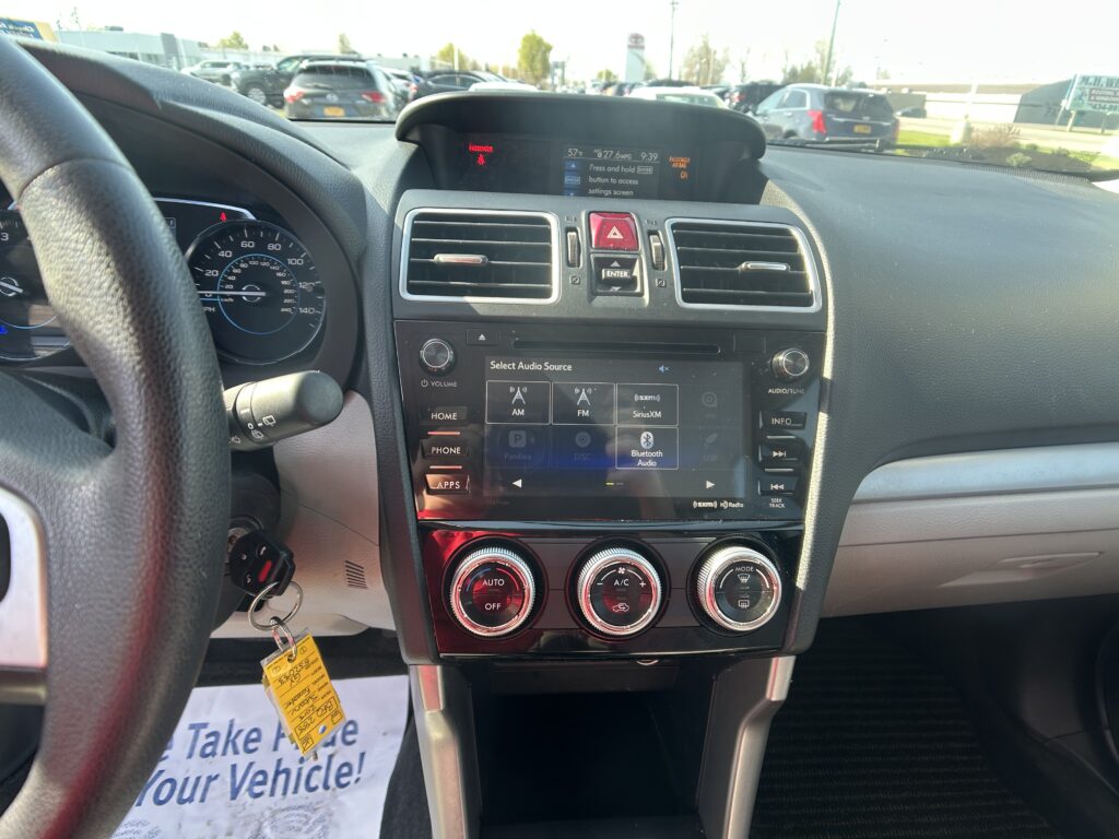 Interior of a car dashboard with a touchscreen infotainment system, circular climate controls, and steering wheel on the left, parked in a lot.