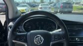 View from driver seat of a Buick, dashboard and steering wheel visible, outside a busy car lot with blue sky.