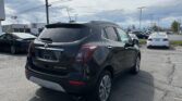 Rear view of a dark gray Buick Encore SUV parked in a dealership lot with other cars and a building in the distance on a sunny day, power lines overhead