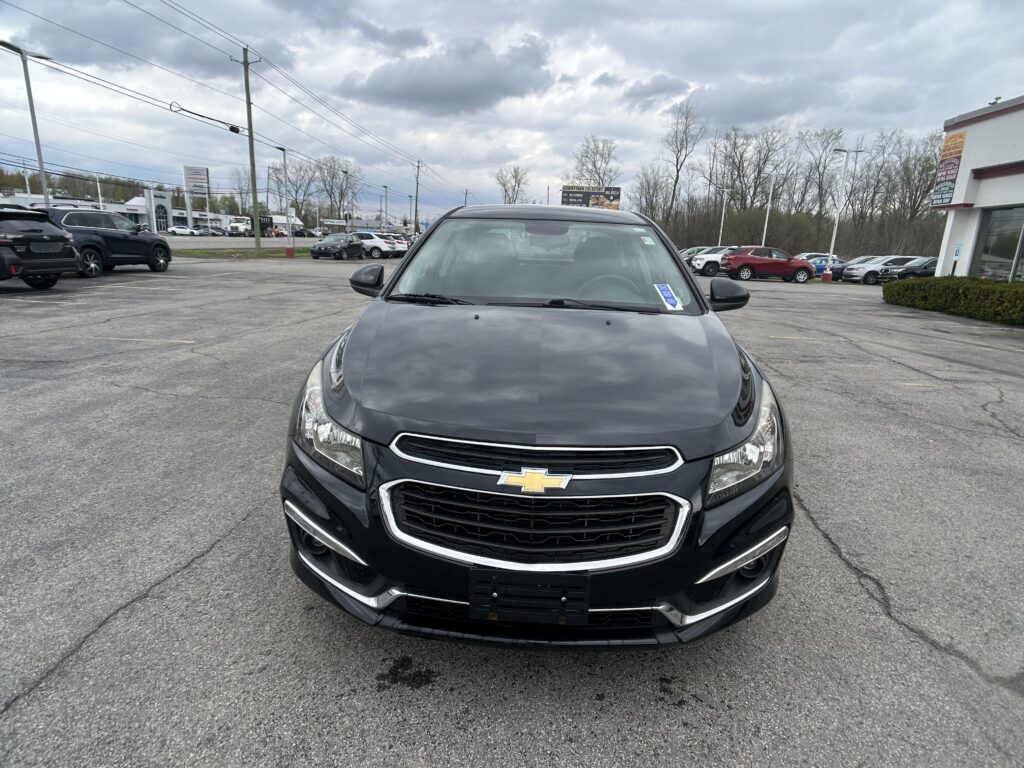 Front view of a dark gray Chevrolet SUV in a parking lot with other cars and a dealership building in the background, under a cloudy sky.