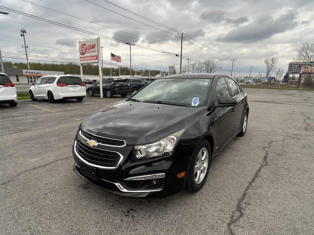 Black Chevrolet SUV in a dealership lot with other vehicles, EZ Loan Auto Sales sign, and an American flag against a cloudy sky.