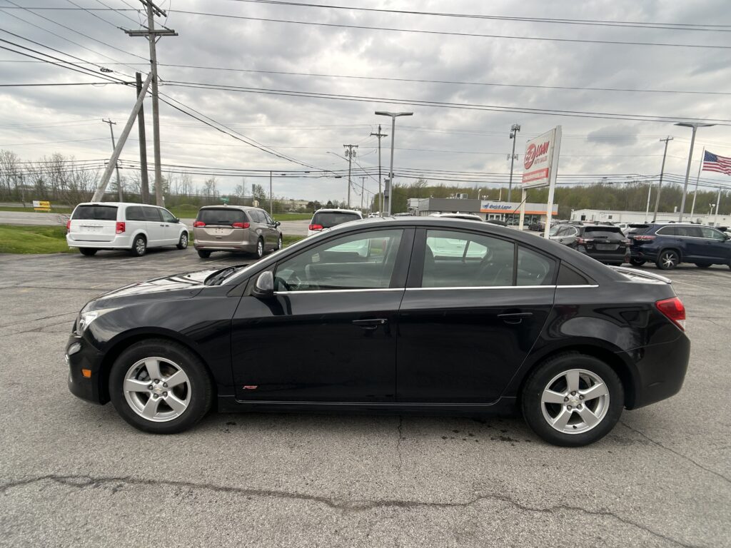 Close-up view of a black sedan in a dealership parking lot, with other cars and a cloudy sky in the background.