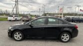 Close-up view of a black sedan in a dealership parking lot, with other cars and a cloudy sky in the background.