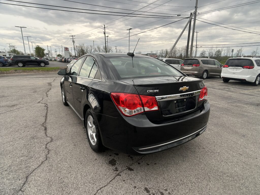 Rear view of a black Chevrolet Cruze in a dealership parking lot with other vehicles and a cloudy sky above.