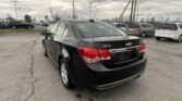 Rear view of a black Chevrolet Cruze in a dealership parking lot with other vehicles and a cloudy sky above.