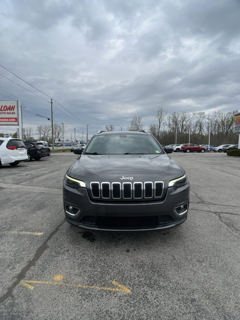 Front view of a gray Jeep SUV parked in a dealership lot under a cloudy sky.