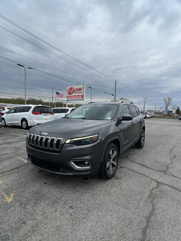 Gray Jeep SUV in a car lot with an E-Z Loan Auto Sales sign in the background and an American flag nearby
