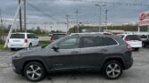 Gray Jeep Cherokee in a dealership parking lot with other cars and overcast sky overhead, power lines in the background.
