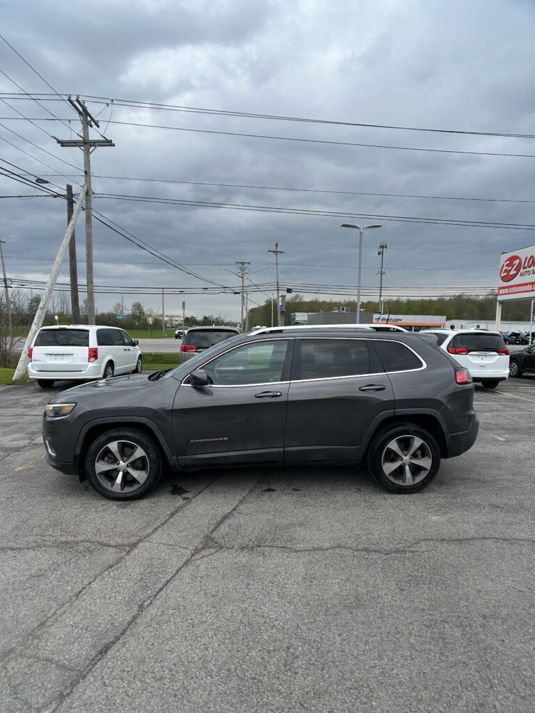 Gray Jeep Cherokee in a dealership parking lot with other cars and overcast sky overhead, power lines in the background.