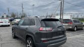 Gray Jeep SUV in a busy outdoor parking lot with multiple cars and a cloudy sky; power lines and utility pole in the background.