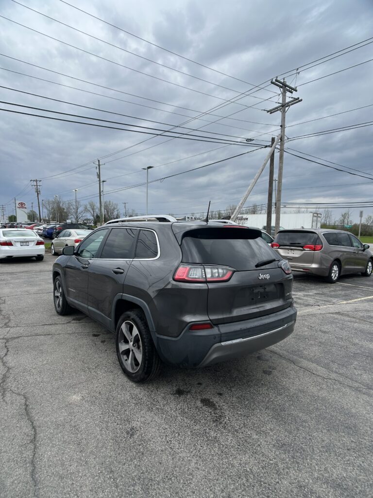 Gray Jeep SUV in a busy outdoor parking lot with multiple cars and a cloudy sky; power lines and utility pole in the background.
