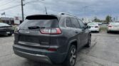 Rear view of a gray Jeep Grand Cherokee Limited in a car dealership lot, with other vehicles and a Toyota dealership building in the background under a cloudy sky.