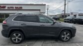 Gray Jeep Cherokee SUV parked in a car dealership lot with an 'Auto Sales' sign and cloudy sky in the background.