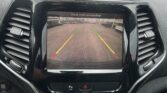 Infotainment screen showing rearview backup camera with guiding lines in a parking lot behind the car, message 'Check entire surroundings' visible on screen.