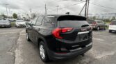 Rear-right view of a black GMC Terrain SUV in a busy car dealership lot, with other cars and utility poles in the background
