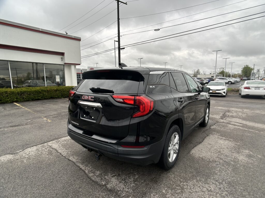 Black GMC Terrain SUV parked in a car dealership lot, building with large windows on the left and other cars in the background under a cloudy sky.