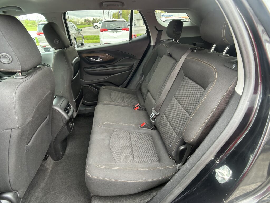 Rear passenger area of a dark fabric car with three seats, headrests, and visible seat belts; outside parking lot seen through windows.