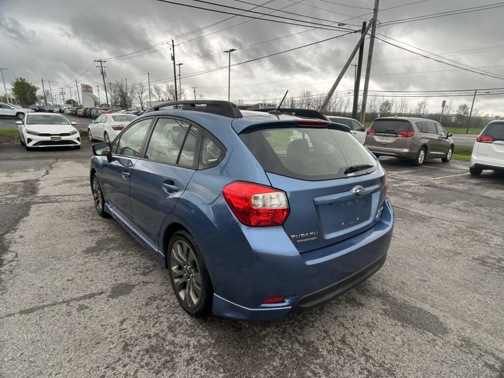 Blue Subaru SUV in foreground, parked in a busy dealership lot on a gray, cloudy day with power lines overhead.