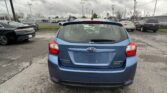 Rear view of a blue Subaru Impreza hatchback in a parking lot, with other cars and a cloudy sky above.