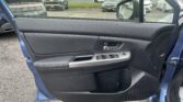 Inside a blue car showing the door panel with window controls, looking out at a car dealership lot with many parked vehicles and a building in the background