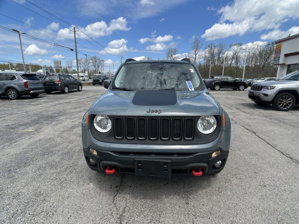 Front view of a gray Jeep with red tow hooks in a busy car dealership lot, with other vehicles and a blue sky above.