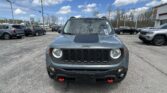 Front view of a gray Jeep with red tow hooks in a busy car dealership lot, with other vehicles and a blue sky above.