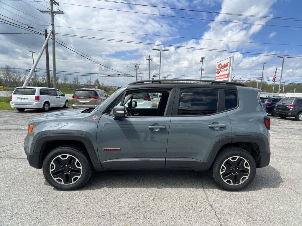 Gray Jeep Renegade SUV in a dealership lot with other cars and power lines overhead, bright blue sky with clouds in the background.