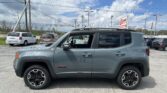 Gray Jeep Renegade SUV in a dealership lot with other cars and power lines overhead, bright blue sky with clouds in the background.