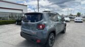 Rear view of a gray Jeep SUV with Trailhawk badge in a dealership lot, white building to the left and power lines overhead.