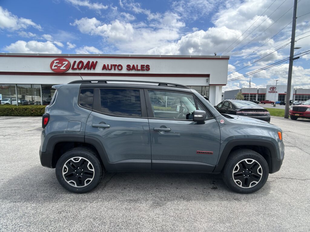 Gray Jeep Renegade SUV in foreground at an E-Z Loan Auto Sales lot under a blue sky with white clouds.