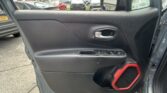 Inside of a car door panel showing a silver door handle, window controls, and a red speaker guard; outside is a dealership parking lot with several trucks and cars.