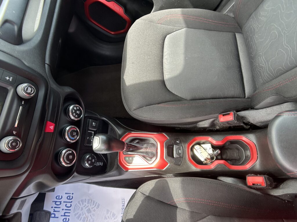 Interior of a car showing a red-accented center console with the gear shifter, climate controls, and red-stitched seats/armrest.