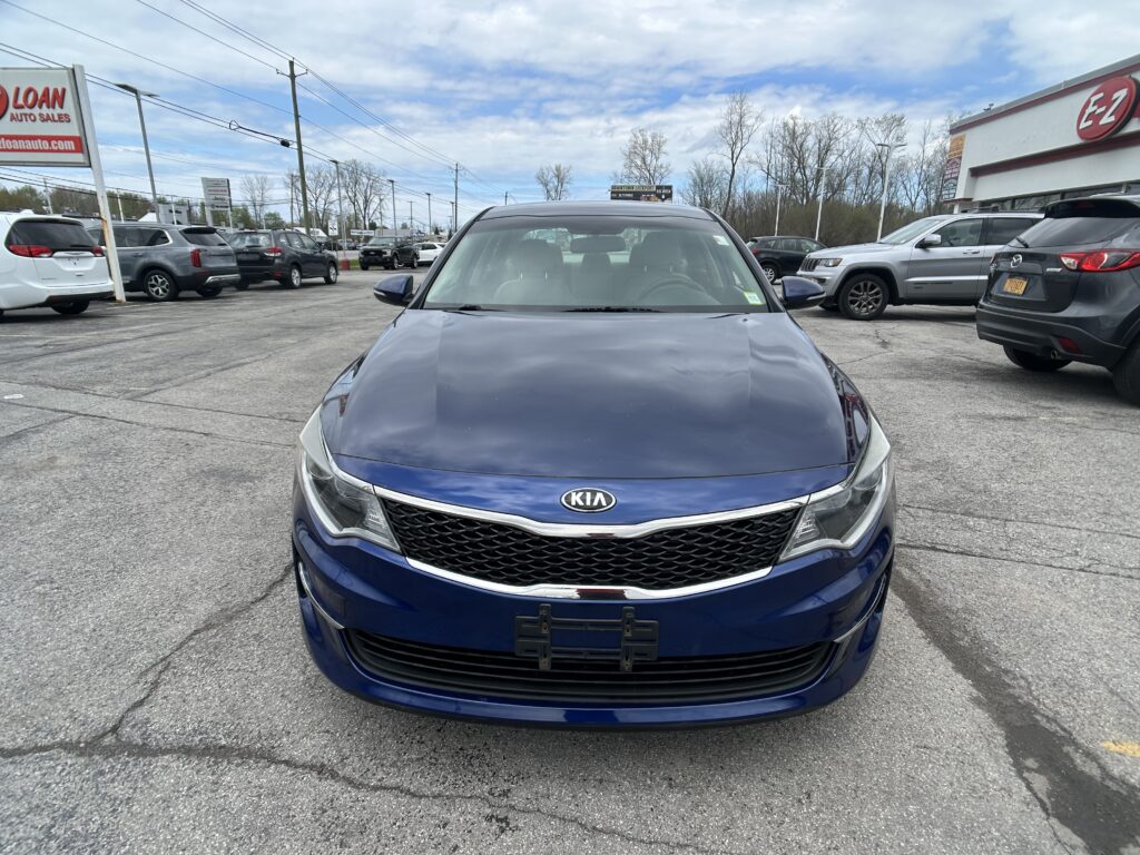 Front view of a blue Kia car in a dealership lot, with other vehicles parked nearby and an EZ dealership sign in the background.