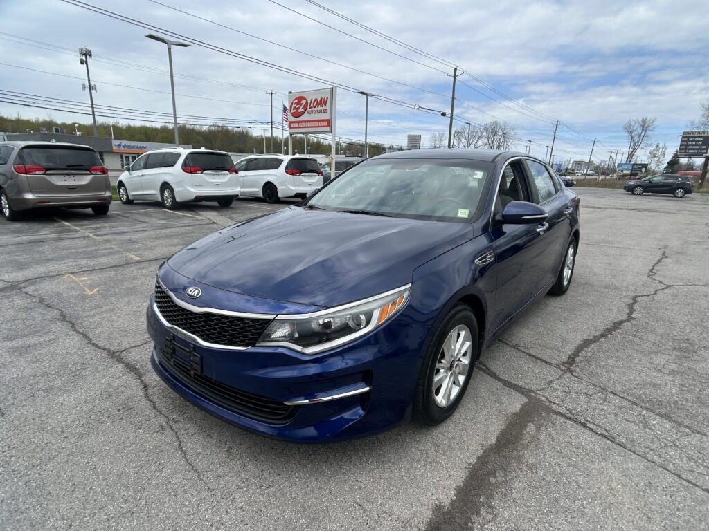 Blue Kia sedan in a parking lot foreground with white minivans and power lines in the background; signs and cloudy sky above.