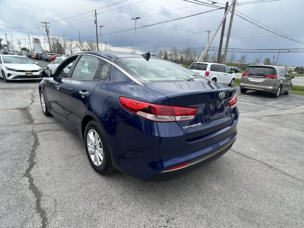 Blue Kia Optima parked in a car dealership lot, with other vehicles and power lines in the background.