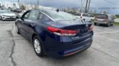 Blue Kia Optima parked in a car dealership lot, with other vehicles and power lines in the background.