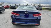 Rear view of a blue Kia Optima in a car dealership parking lot with other vehicles and dealership buildings in the background under a cloudy sky.