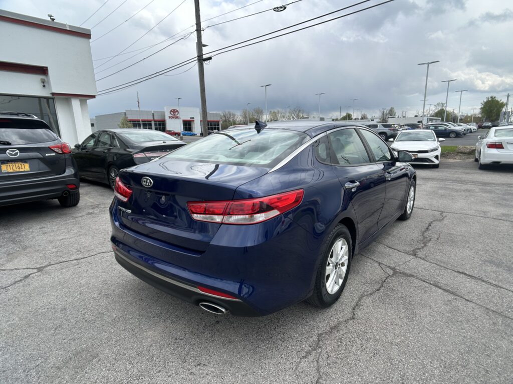 Back of a navy blue Kia sedan in a car dealership lot with many other cars and a Toyota building in the background.