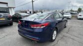Back of a navy blue Kia sedan in a car dealership lot with many other cars and a Toyota building in the background.